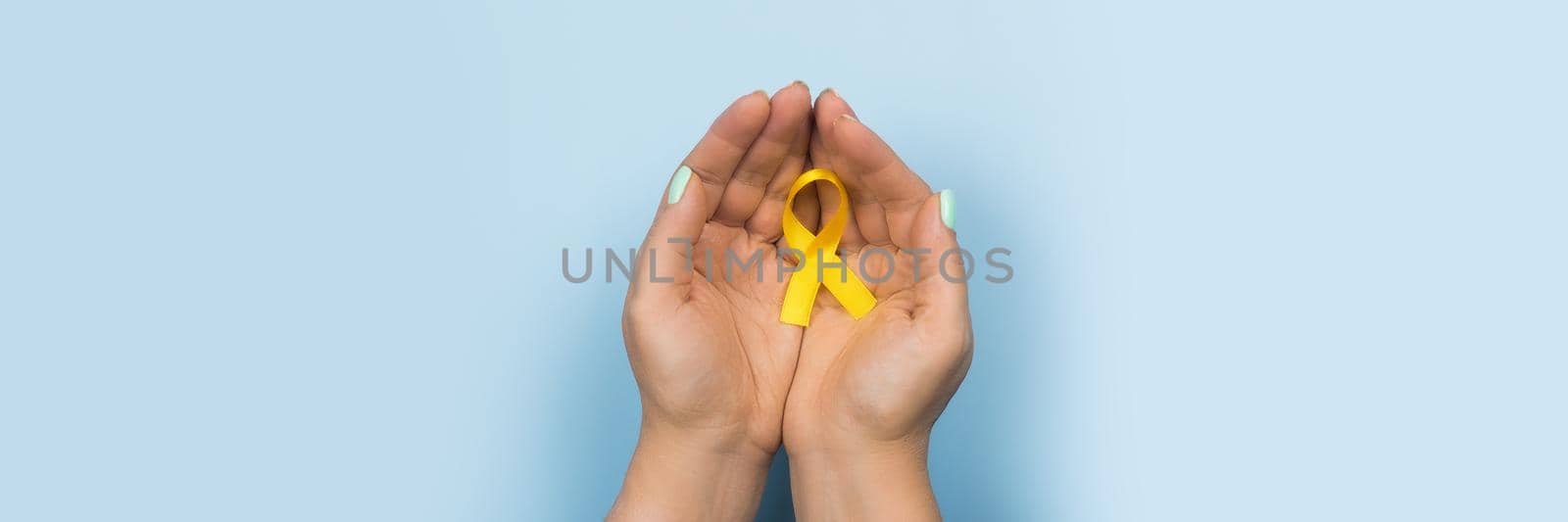 Yellow gold ribbon in the hands of a girl on a blue background. Children cancer concept. Awareness month. Childhood Cancer Day February, 15. Web banner.