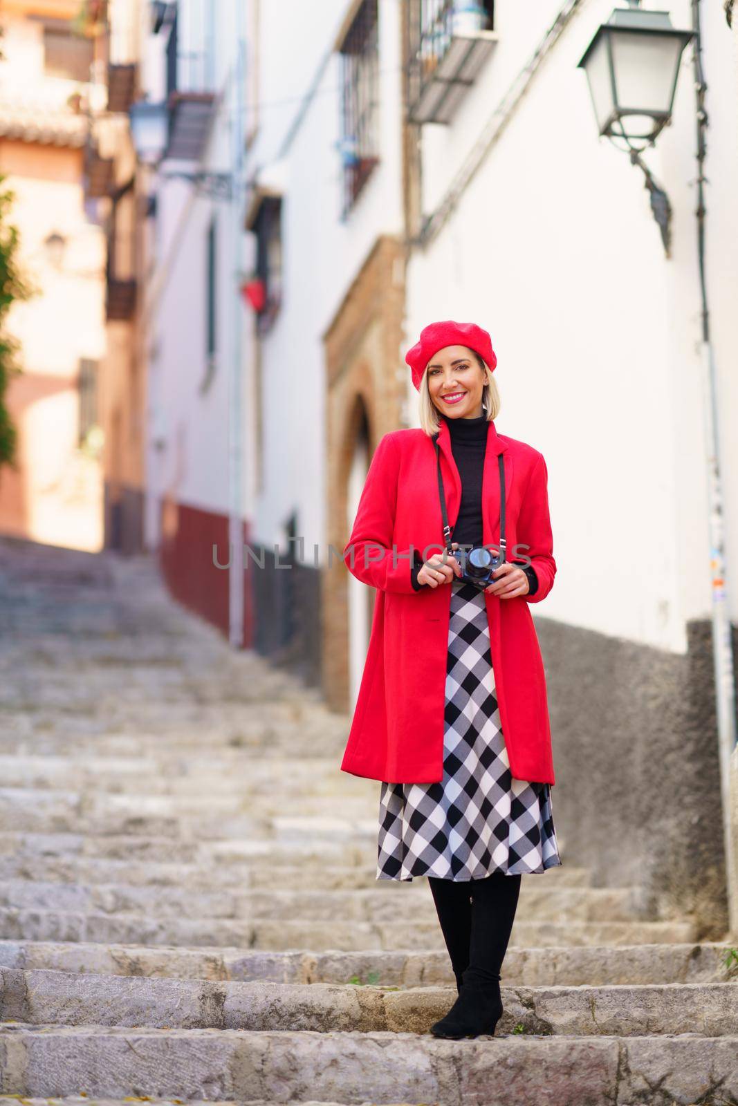 Full body of delighted young female traveler in elegant red coat, checkered skirt and beret smiling and looking at camera while standing on stairs in old town