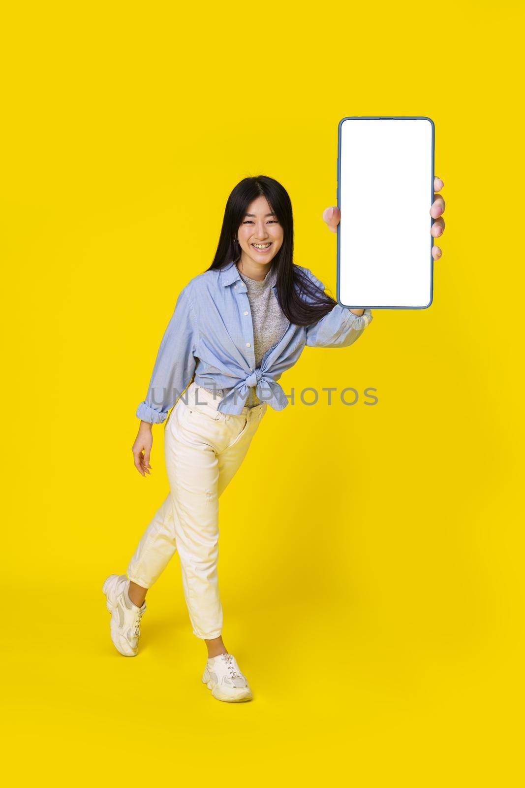 Charming young asian girl holding smartphone showing white screen mobile app advertisement and excited smile on camera wearing casual isolated on yellow background. Product placement by LipikStockMedia