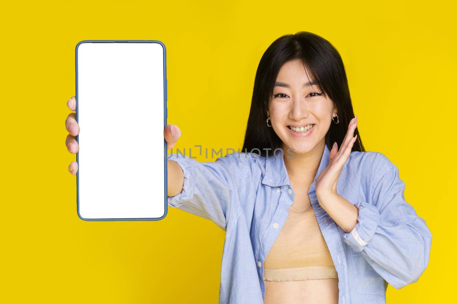 Amazed young asian girl showing mobile phone white white screen and excited smile on camera isolated on yellow background. Girl using smartphone. Selective focus on girl by LipikStockMedia