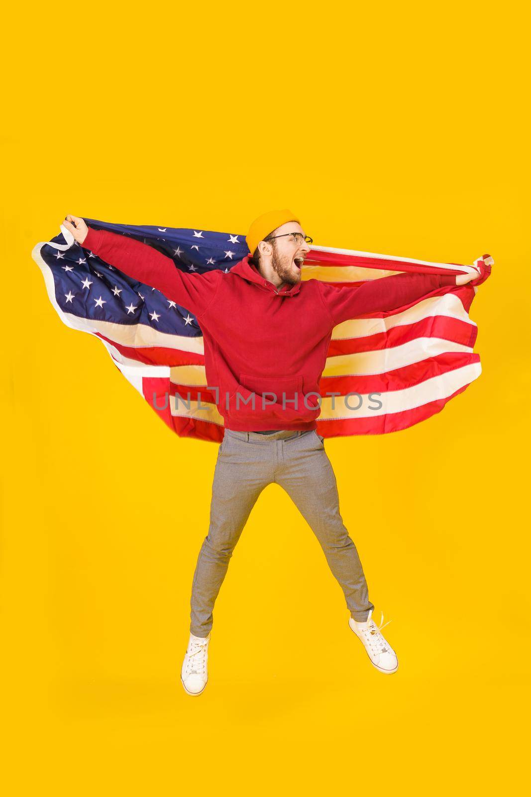 Happy 4th july, handsome young man celebrates jumping hight with USA flag behind his back isolated on yellow background. Freedom is in your life. Cheerful young man with American flag.