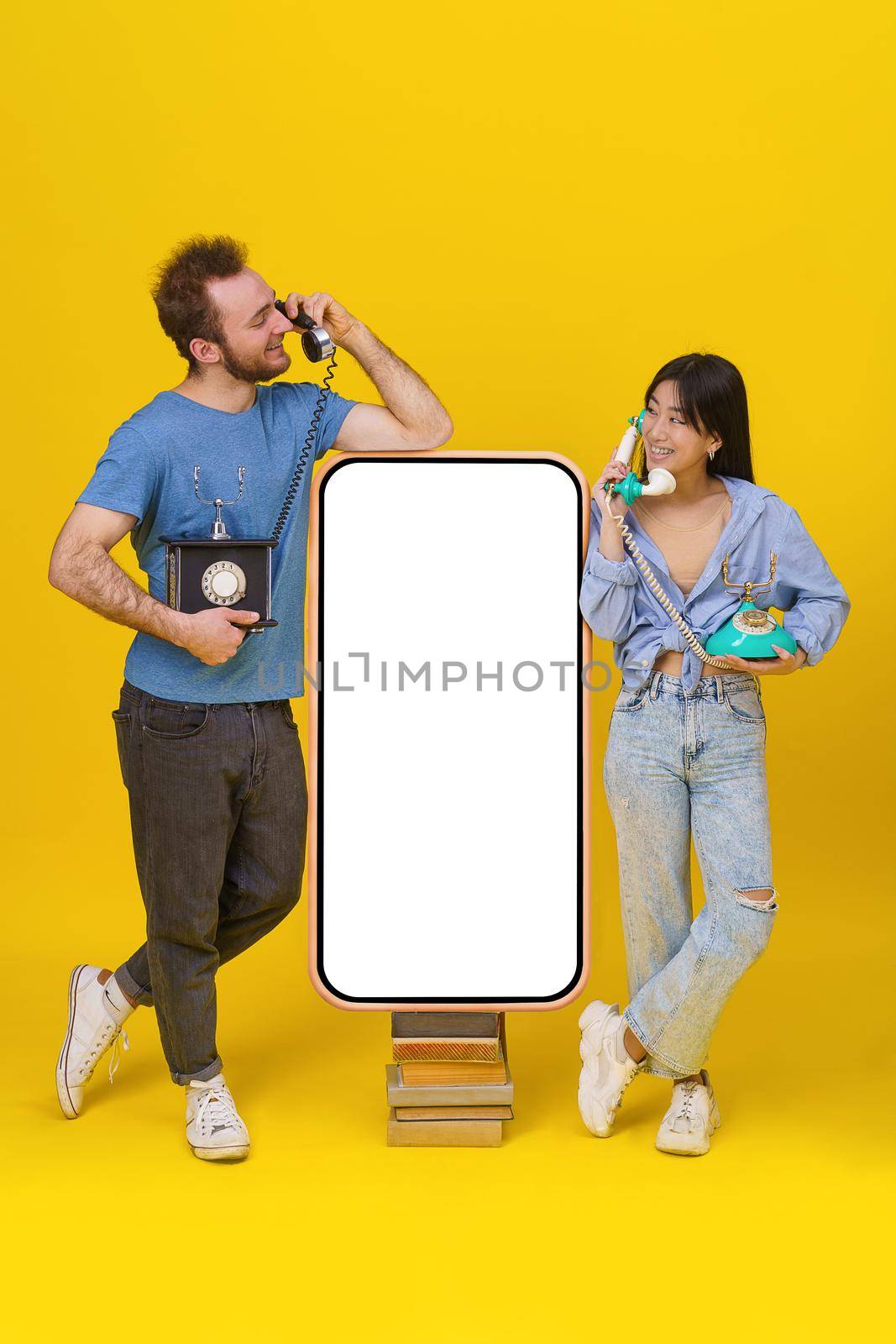 Love talks, handsome guy and asian girl using vintage phones leaned on huge smartphone on old books with blank screen, happy smiling isolated on yellow background. Mock up, product placement.