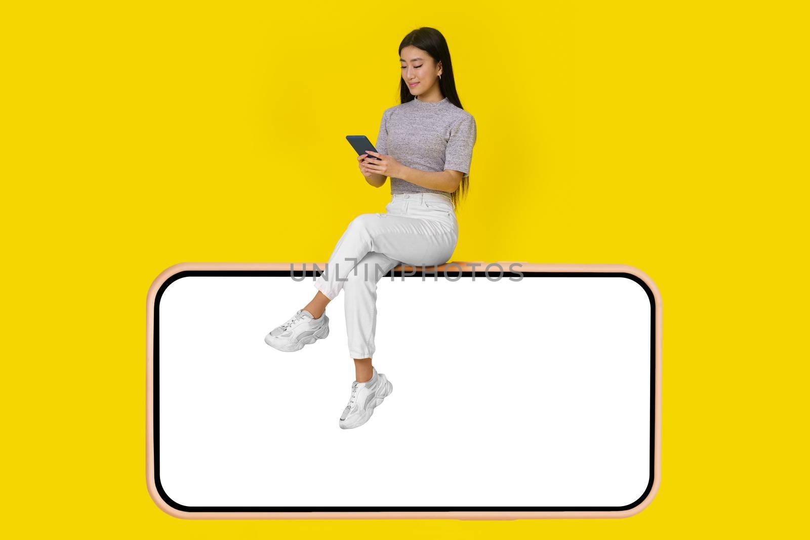 Asian girl with phone in hands sit on huge smartphone with white screen wearing casual white jeans isolated on yellow background. Mock up product placement mobile app advertisement. Copy space.