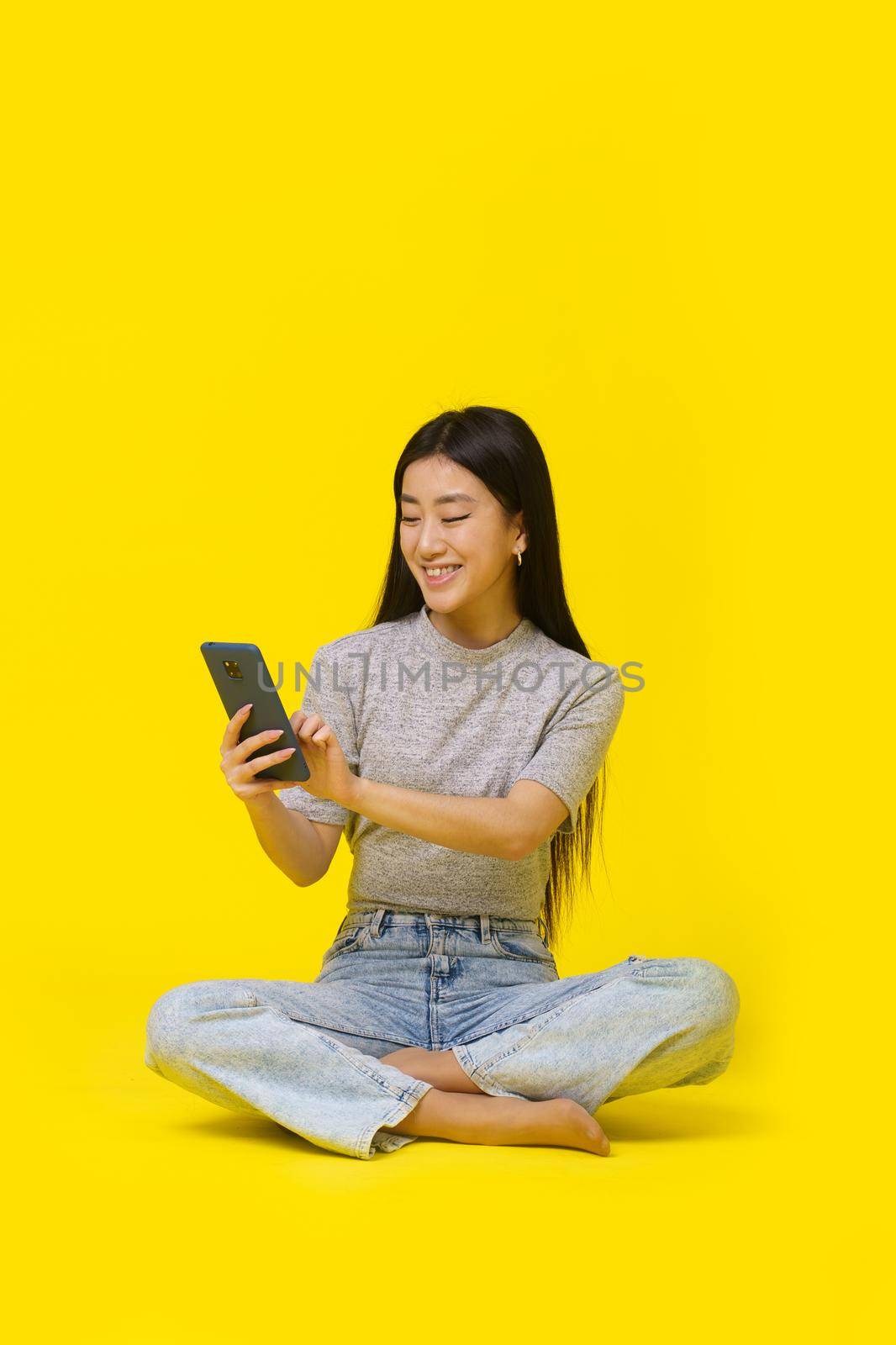 Asian young girl on the floor with phone in hands texting or shopping online playing game isolated on yellow background. Mobile app advertisement product placement. Copy space by LipikStockMedia