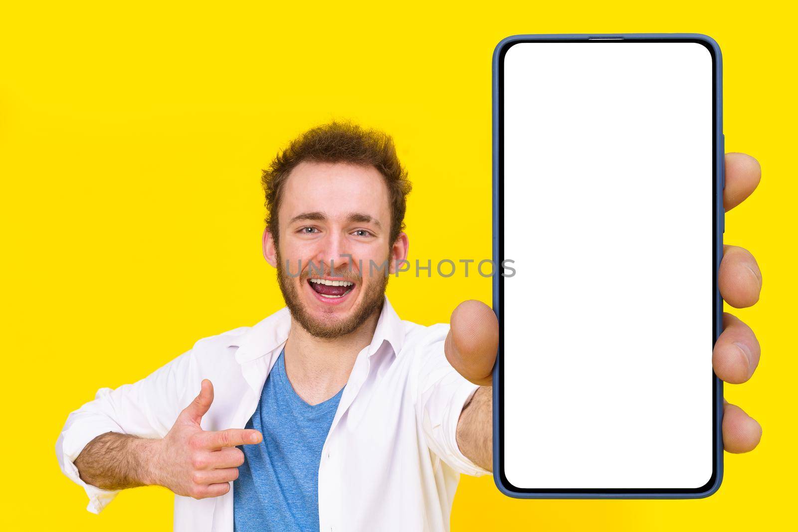 Check this app young man pointing at smartphone showing a white empty screen game, bet, lottery win isolated over yellow background. Product placement for mobile application advertisement by LipikStockMedia