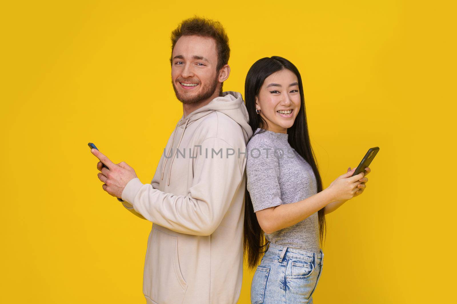Caucasian guy and asian girl standing back to back smiling holding smartphones in hand looking at camera mobile app advertisement isolated on yellow background. Product placement.