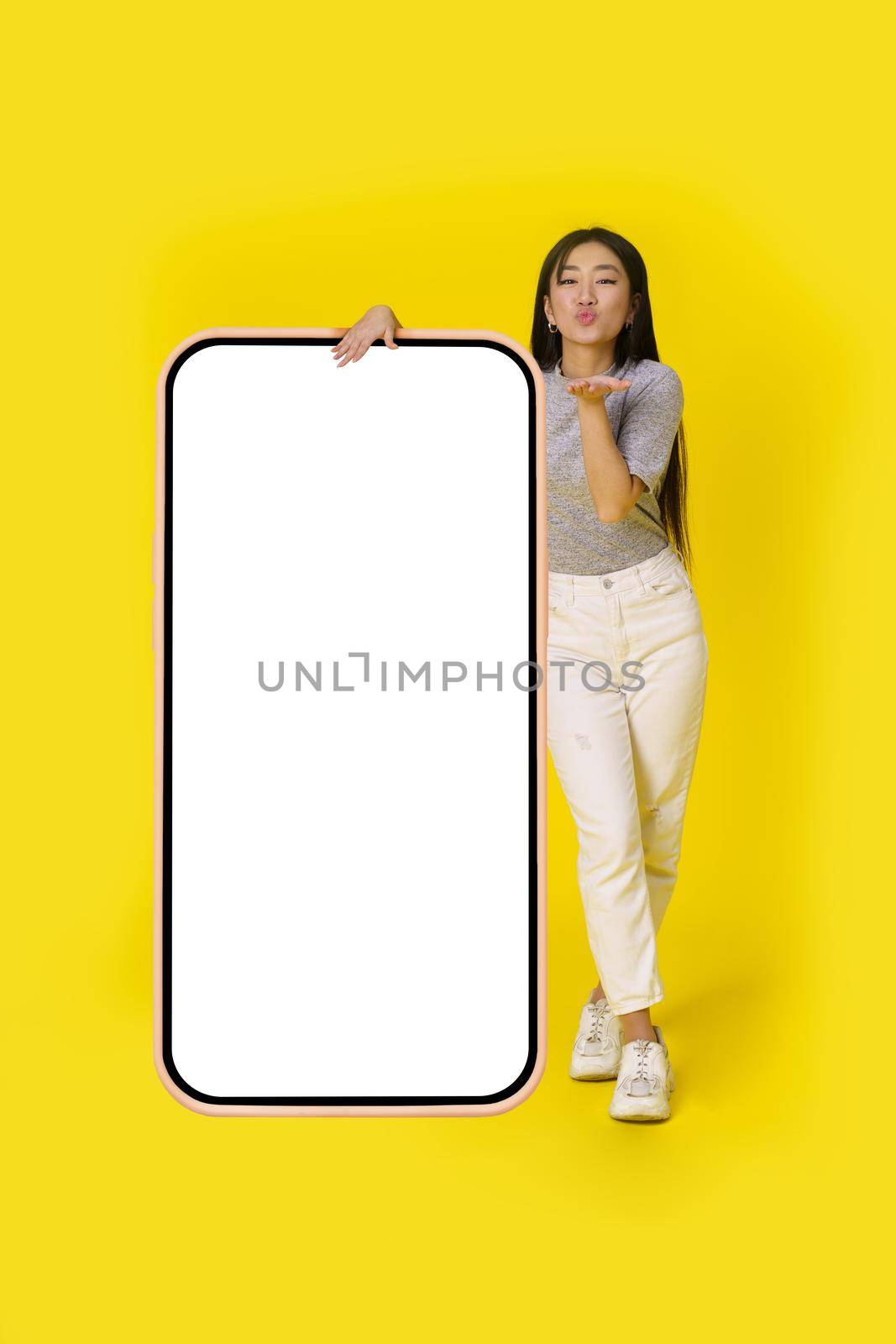 Sending air kisses asian girl stand near huge, giant smartphone with white screen dressed in casual isolated on yellow background. Free space mock up mobile app advertising by LipikStockMedia