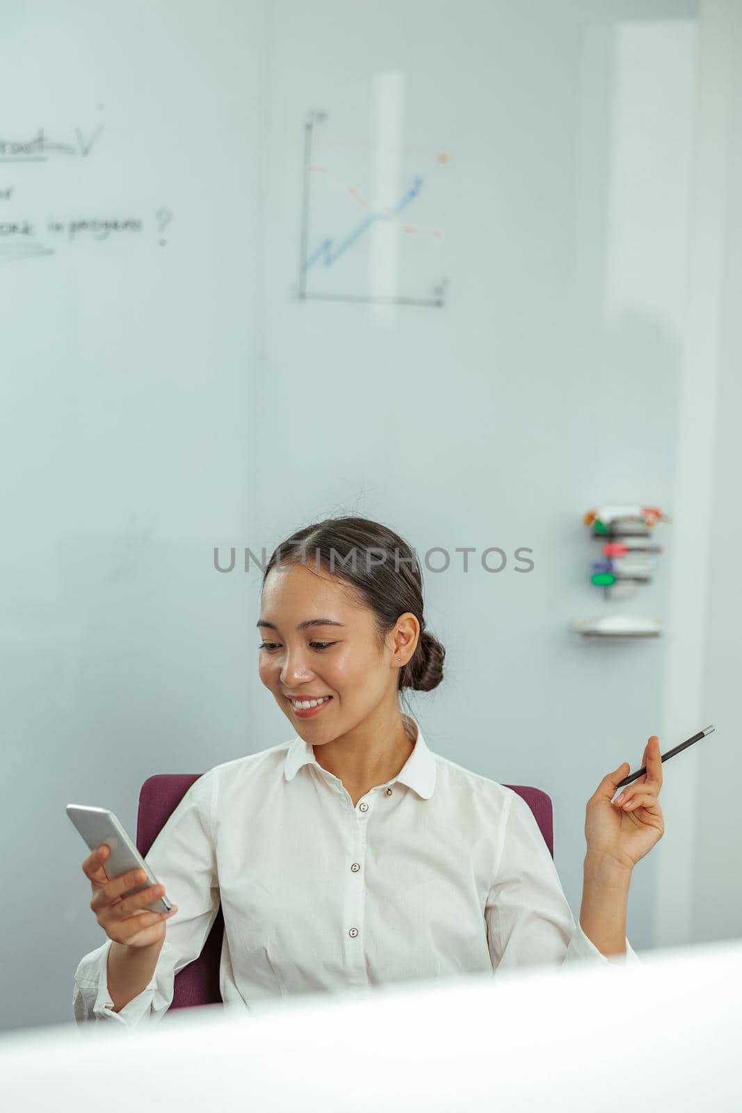 Smiling asian woman worker looking phone while sitting on her workplace in office by Yaroslav_astakhov