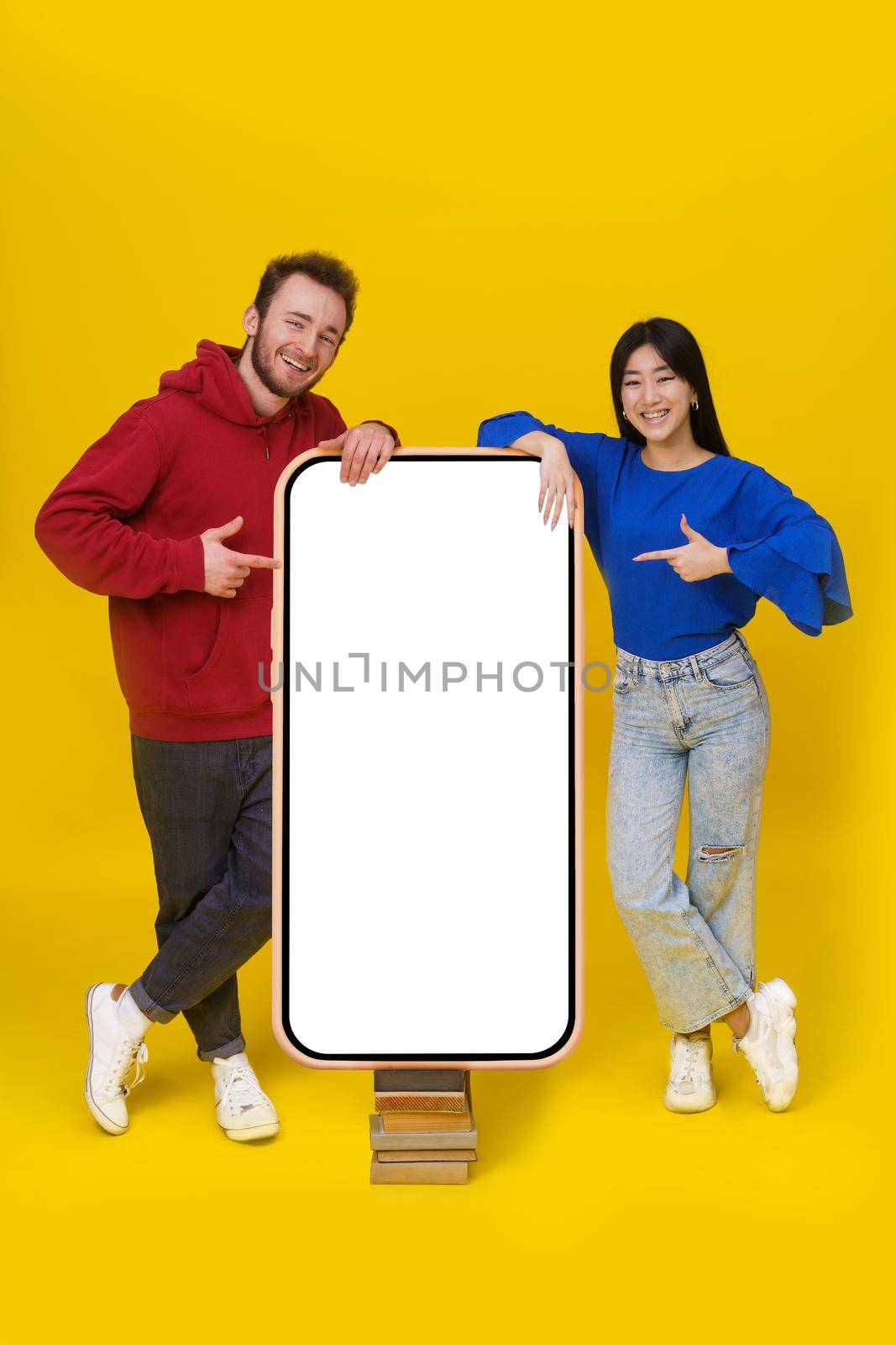 Caucasian guy and asian girl leaned on huge, giant smartphone standing on old books with white screen pointing at it looking at camera mobile app advertisement isolated on yellow background.