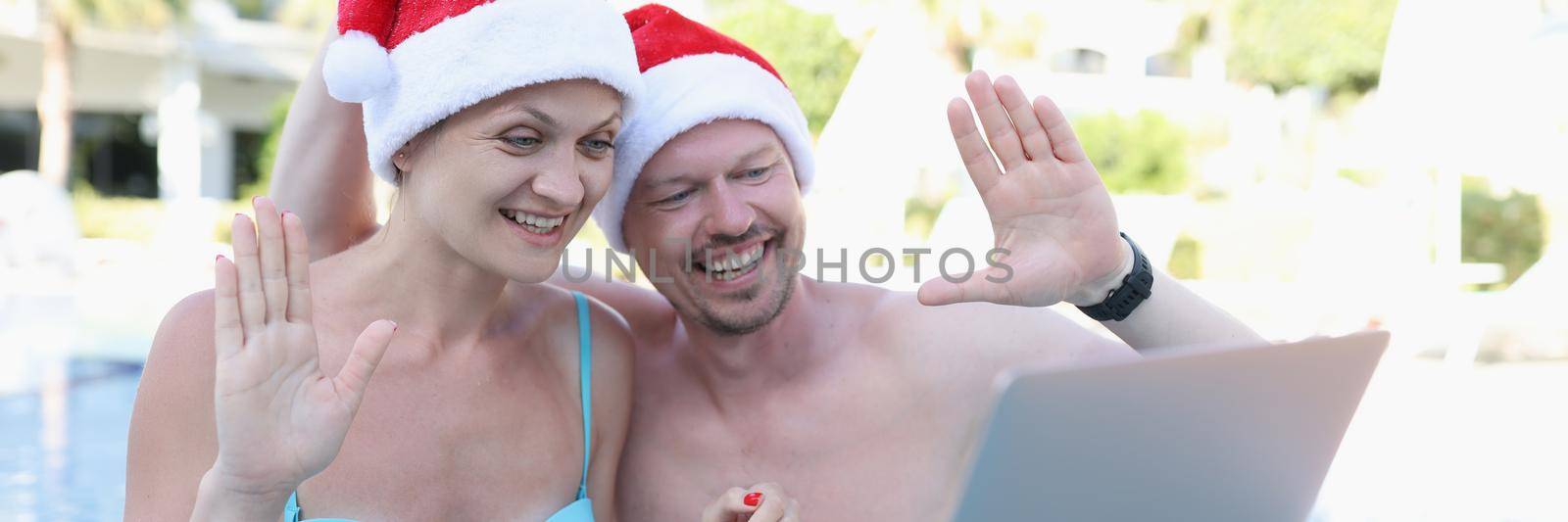 A man and a woman in santa claus hats in the pool use a laptop, close-up. Video conference, remote access