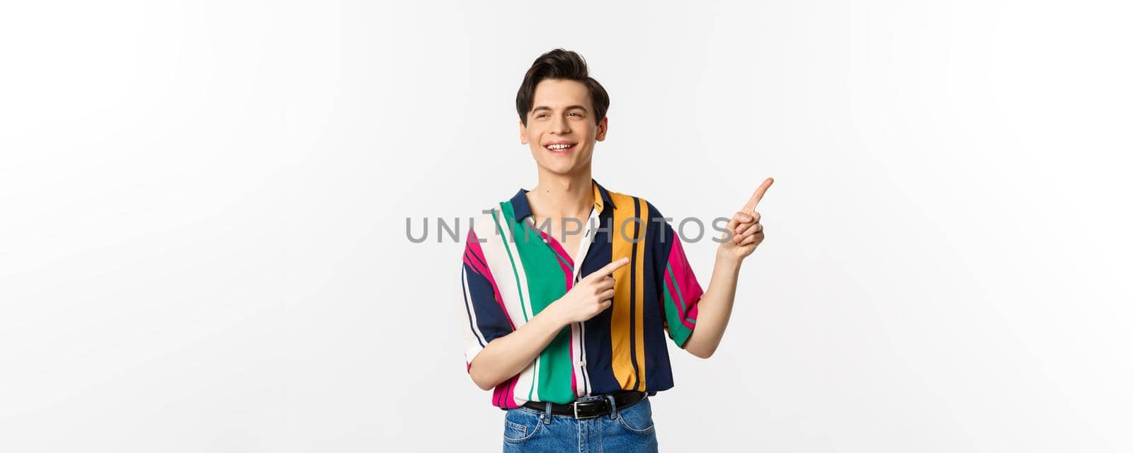 Carefree guy in stylish shirt pointing fingers at upper right corner, showing promo banner or logo, smiling and talking to someone, standing over white background by Benzoix
