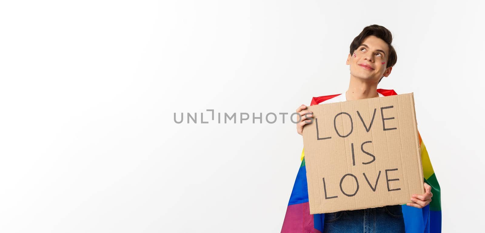 Dreamy young queer person smiling and looking at upper left corner, holding love is love sign for pride parade, wearing Rainbow flag, white background by Benzoix