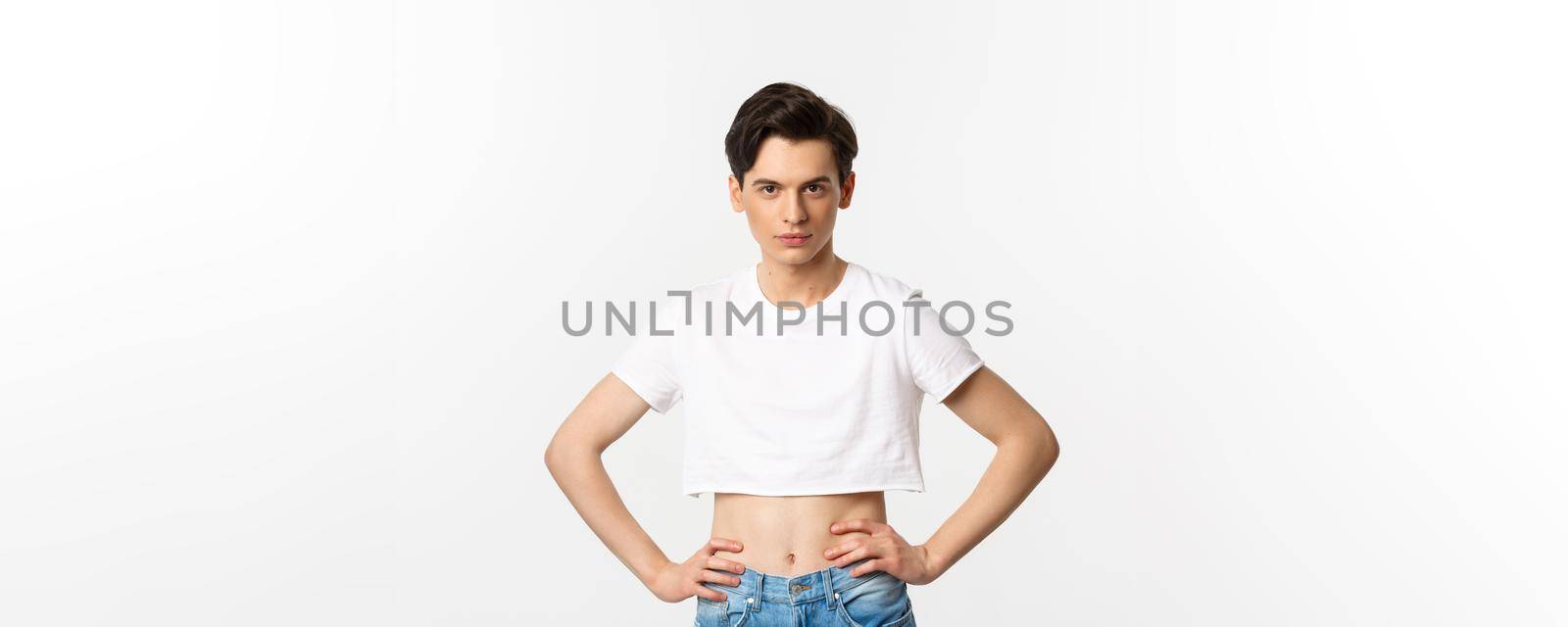 Handsome androgynous man in crop top looking confident at camera, holding hands on waist. Lgbtq activist standing over white background with sassy expression.