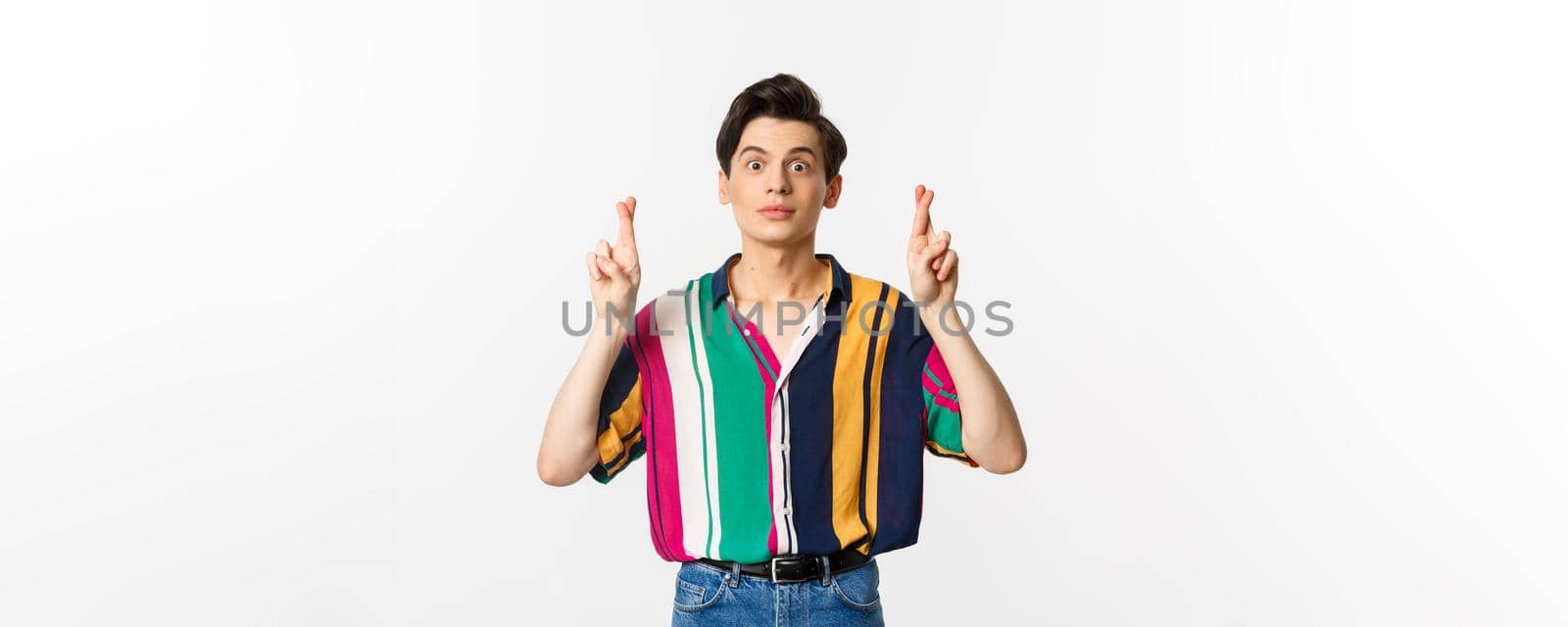 Hopeful young androgynous man cross fingers for good luck, making wish, waiting for something, standing over white background.