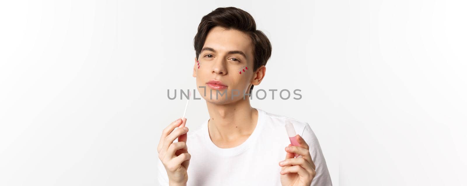Close-up of beautiful queer person with glitter on face applying lips gloss and looking self-assured at camera, standing over white background.