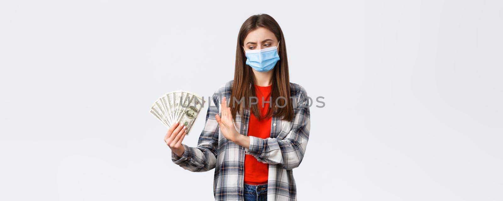 Money transfer, investment, covid-19 pandemic and working from home concept. Displeased girl in medical mask rejecting taking cash as bribe, show stop sign to dollars and frowning bothered.