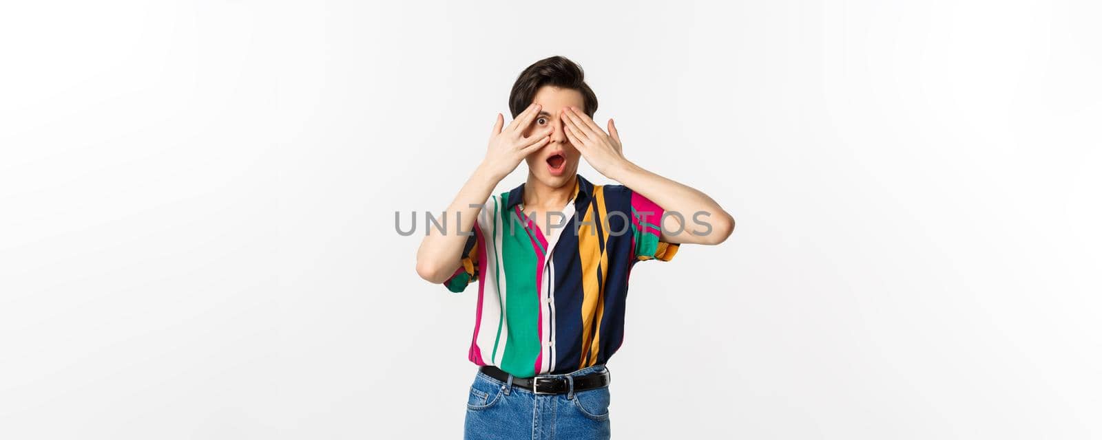 Image of handsome young gay man open eyes to see surprise, peeking through fingers with amazed emotion, standing over white background.