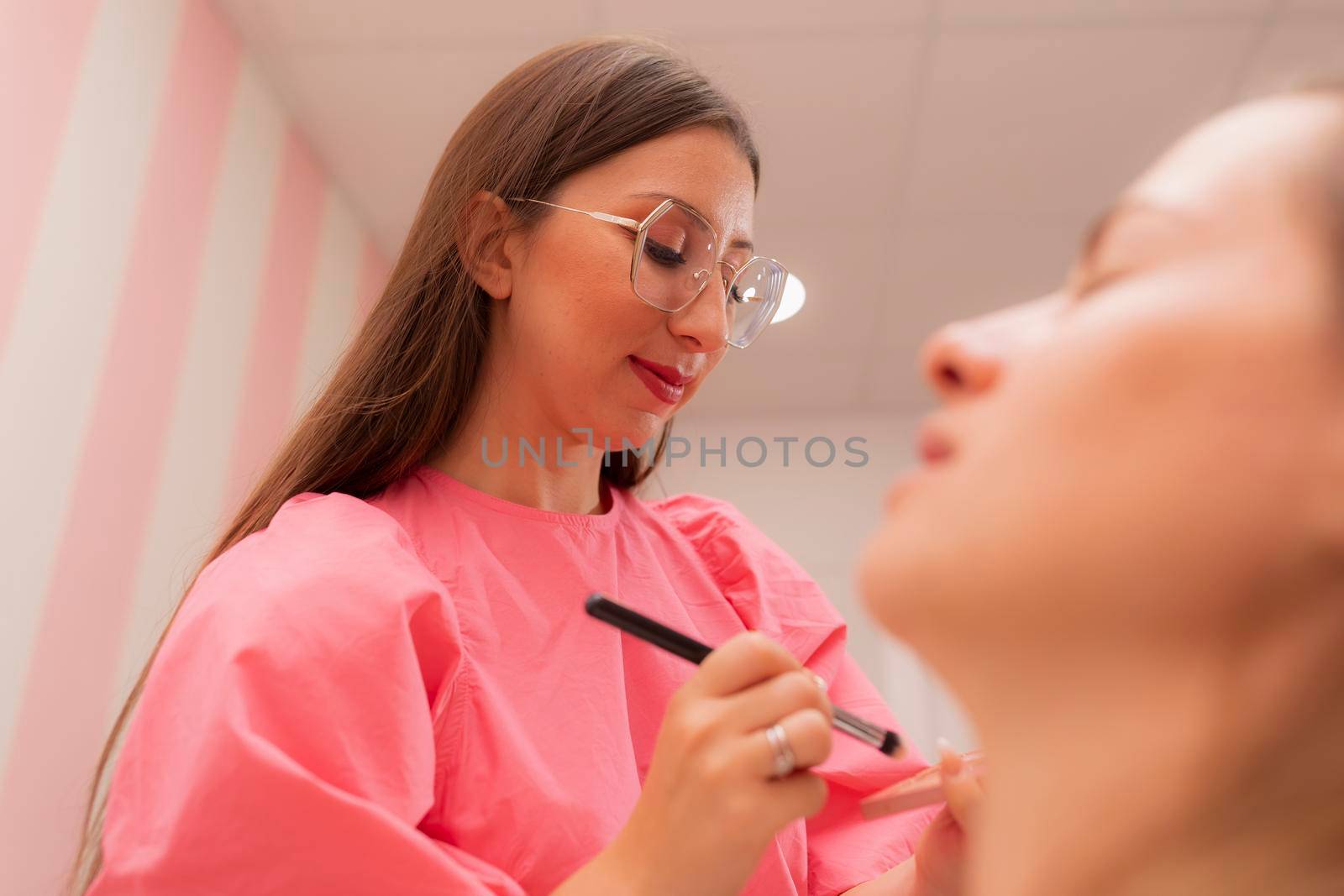 A beautician woman is brushing the foundation on the palette she is holding to apply it onto the customer's face at the beauty salon