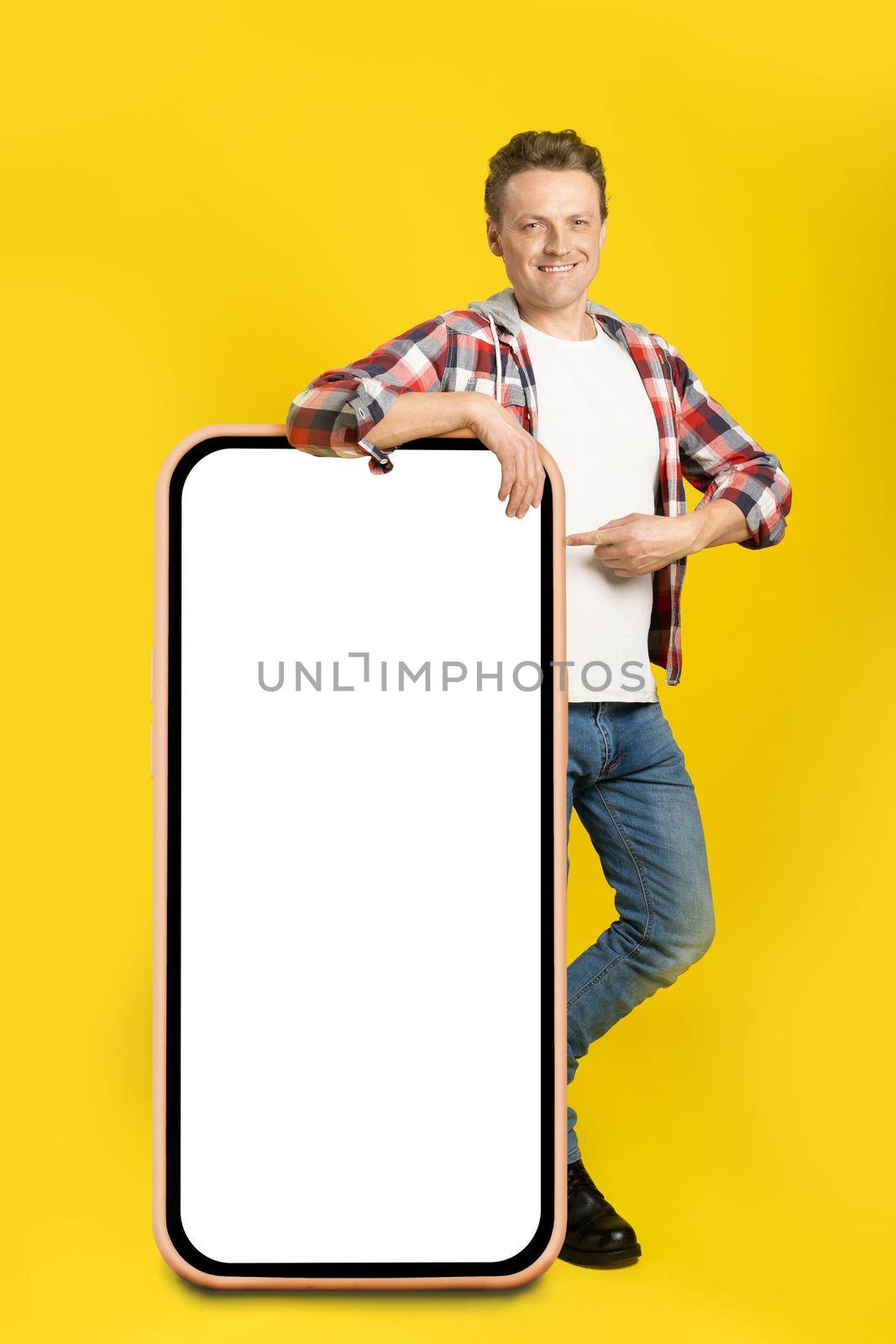 Pointing finger handsome caucasian man leaned on giant, huge smartphone with blank white screen, wearing casual outfit isolated on yellow background. Free space mock up by LipikStockMedia