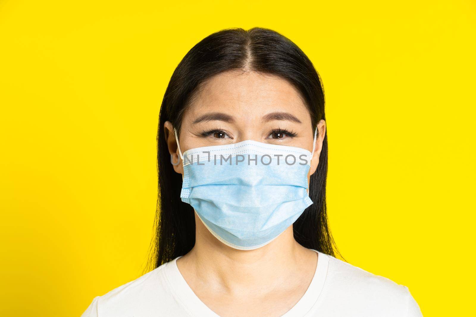 Happy, safe of pandemic mature asian woman wearing medical face mask coronavirus or monkeypox prevention. Charming middle age woman in white t-shirt and medical mask on yellow background by LipikStockMedia