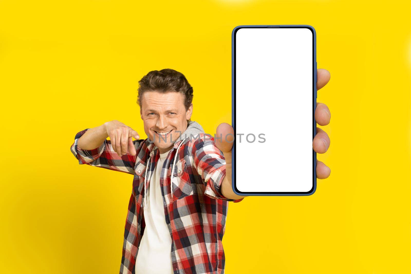 Yo, look at this. Happy handsome man pointing at huge smartphone with white empty screen, wearing red plaid shirt, cellphone display mock up isolated on yellow background. Mobile app advertisement by LipikStockMedia