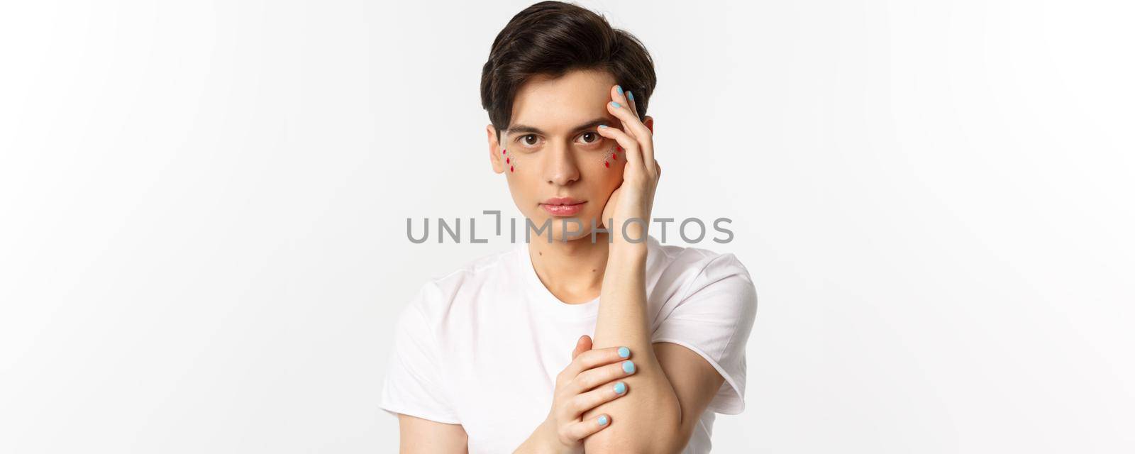 People, lgbtq and beauty concept. Close-up of beautiful queer man touching face with fingers with blue nail polish, standing over white background.