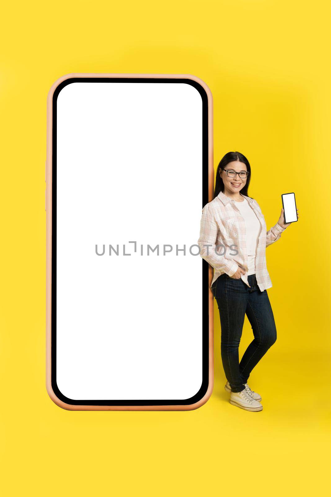 Charming middle aged asian woman with phone in hands showing white screen leaned back on giant, huge smartphone with white screen wearing casual isolated on yellow background. Free space mock up by LipikStockMedia