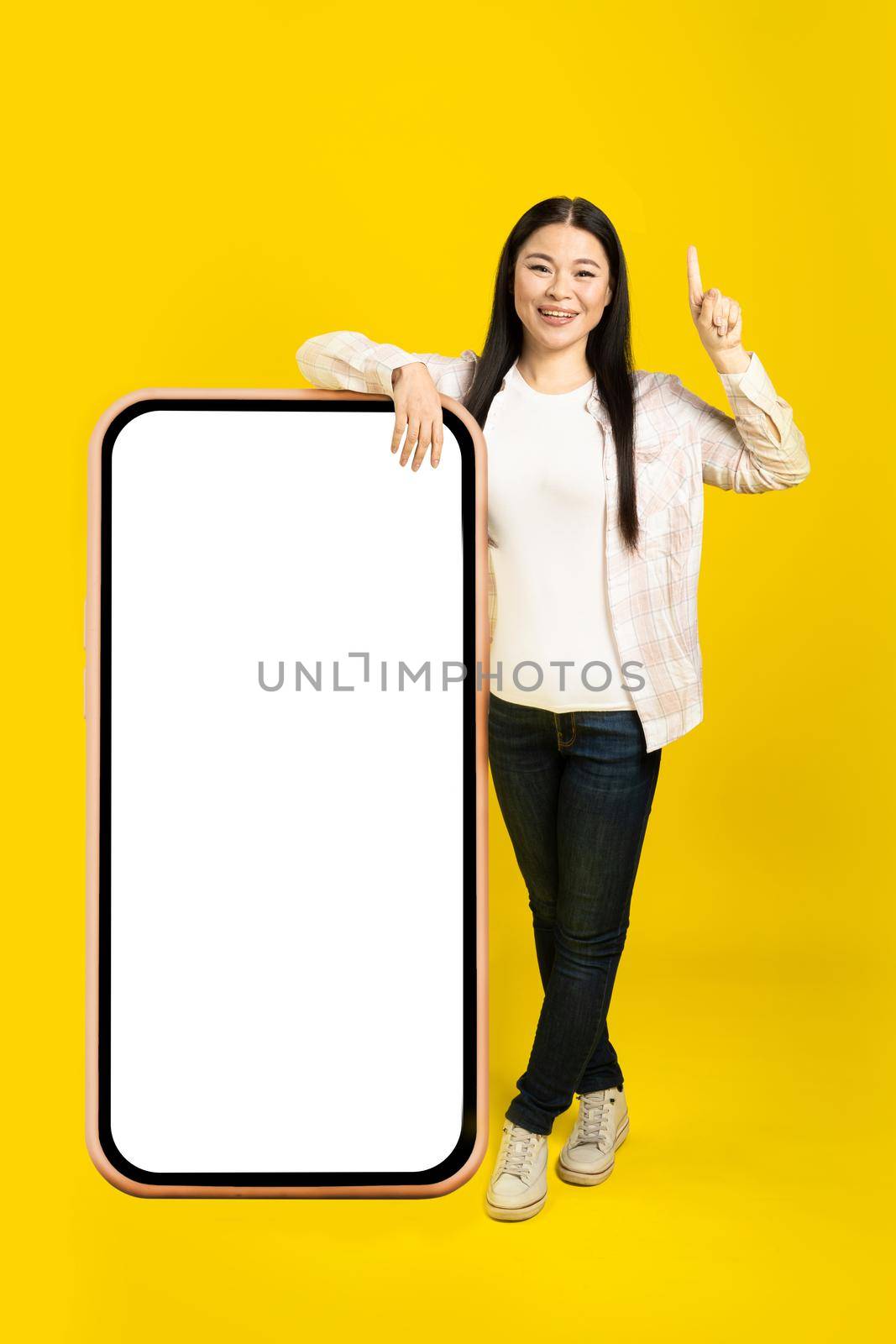 Pointing finger up middle age asian woman leaned on giant, huge smartphone with blank white screen, wearing casual outfit isolated on yellow background. Free space mock up.