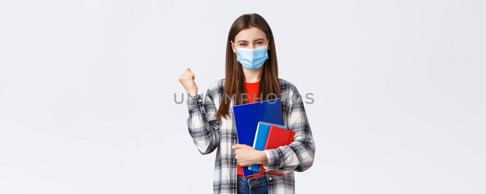 Coronavirus pandemic, covid-19 education, and back to school concept. Rejoicing, cheerful female student in medical mask happy entering cool university, fist pump in success, holding notebooks by Benzoix