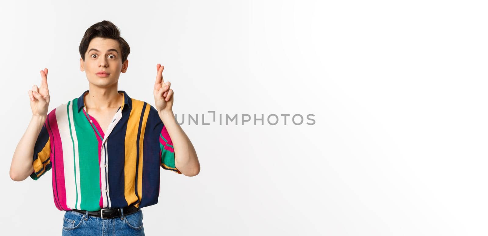 Hopeful young androgynous man cross fingers for good luck, making wish, waiting for something, standing over white background.