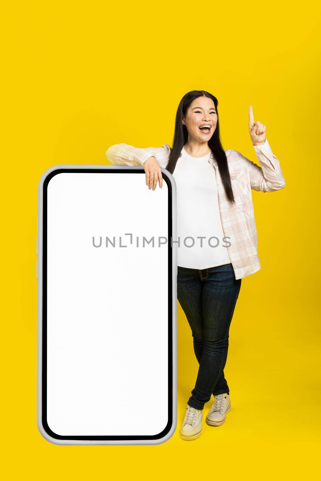 Middle age asian woman excited pointing finger up leaned on giant, huge smartphone with blank white screen, wearing casual outfit isolated on yellow background. Free space mock up by LipikStockMedia