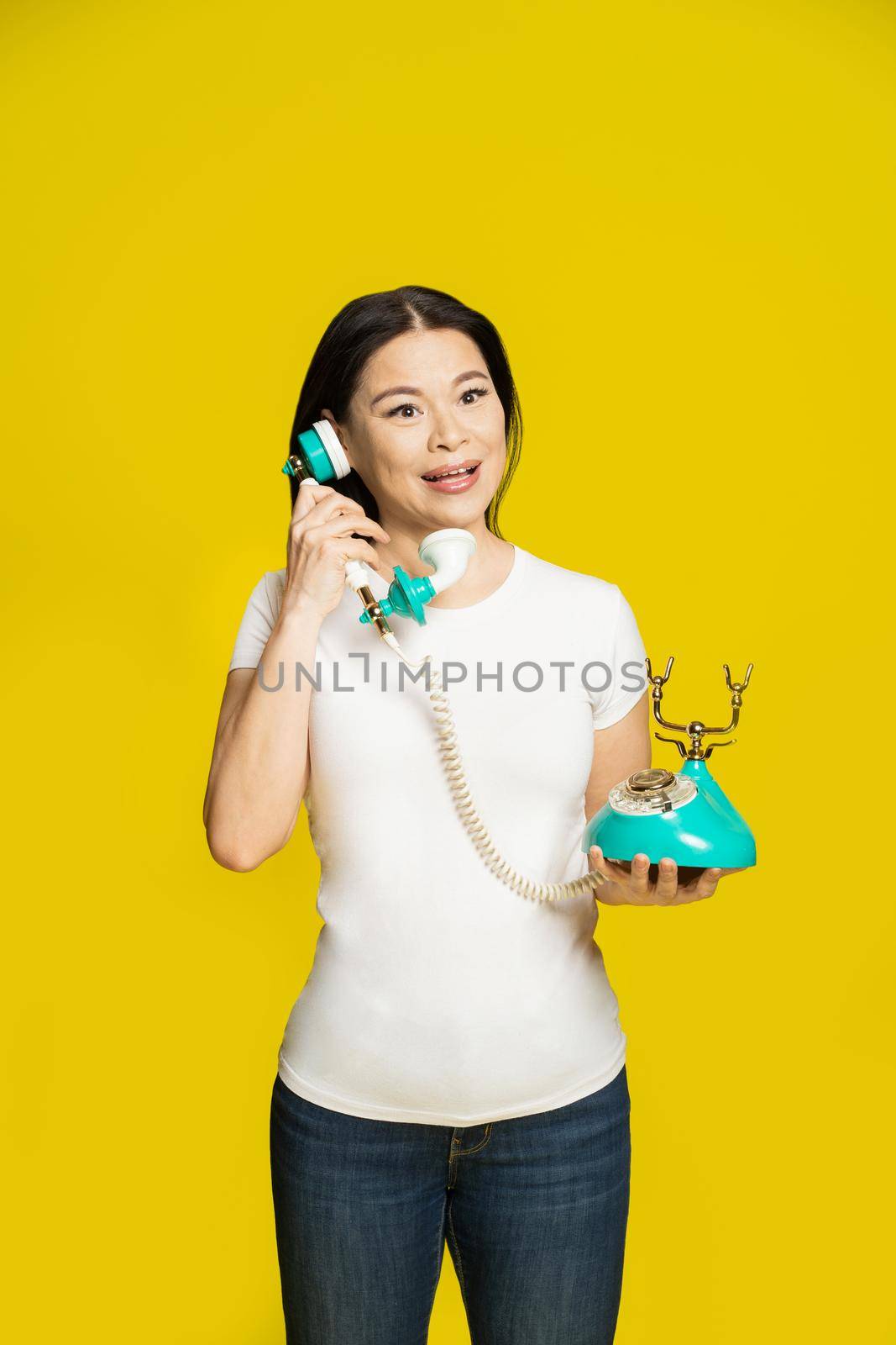 Charming funny asian woman using vintage, retro telephone in hands wearing white t-shirt isolated on yellow background. Excited asian woman with retro phone. Communication concept by LipikStockMedia