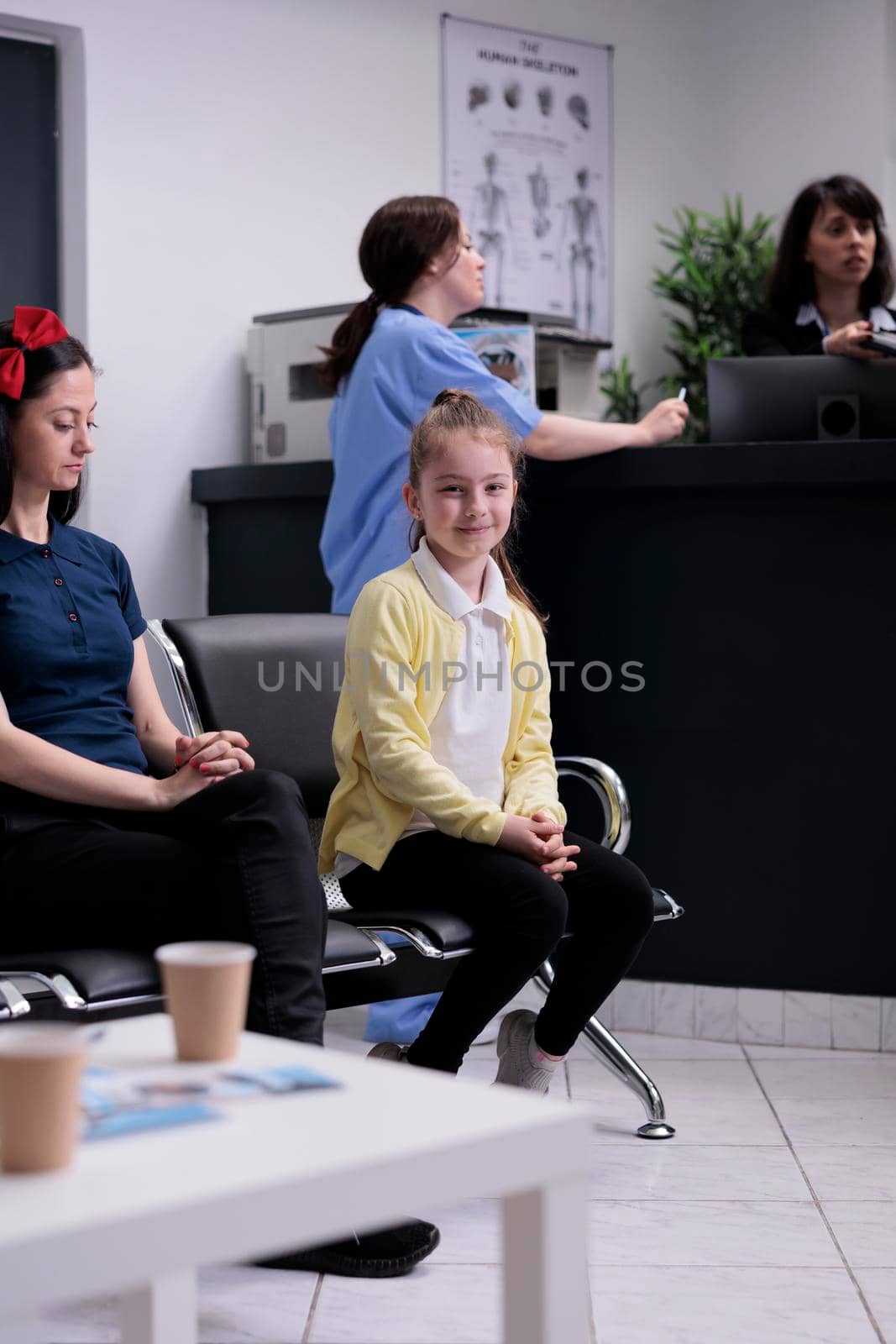 Portrait of smiling young girl with her mother sitting in hospital lobby waiting to see pediatric doctor. Parent with child at private clinic reception front desk visiting pediatrician specialist. by DCStudio