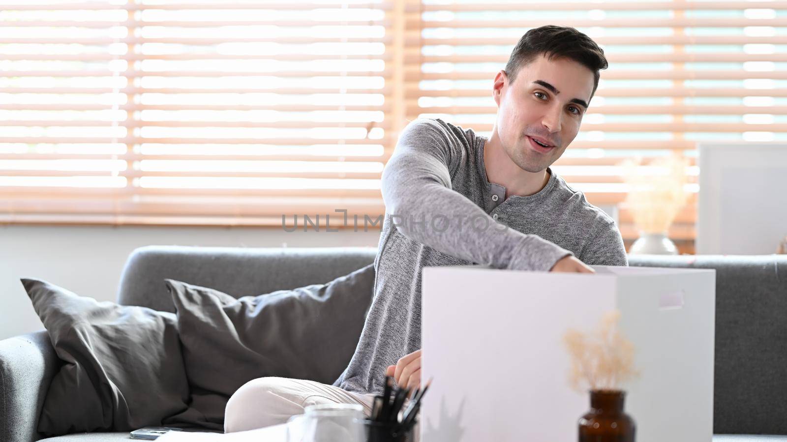 Young man sitting on couch and packing belongings into carton containers preparing for moving into new house.