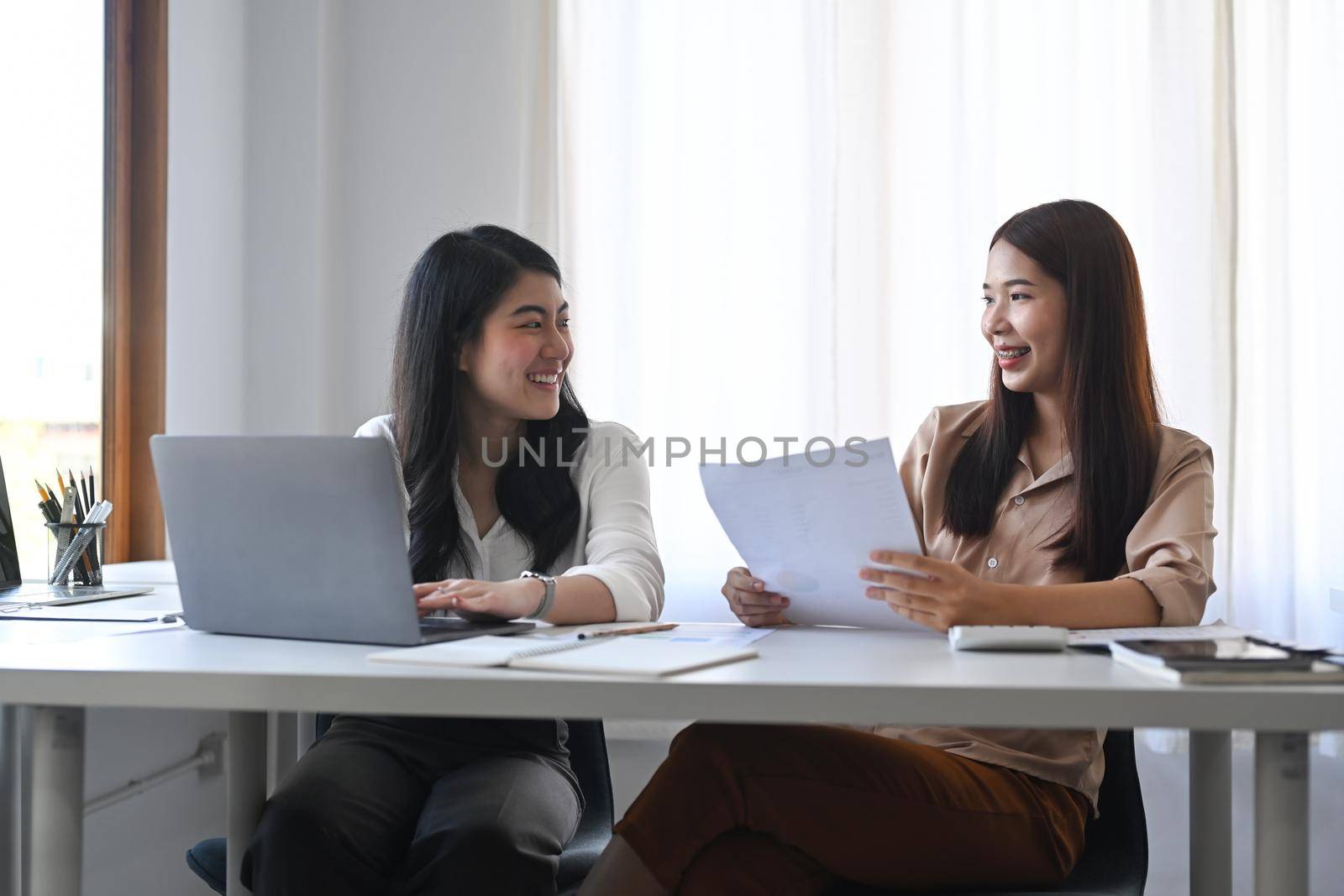 Two happy businesswomen sharing ideas, discussing project strategy together at office.