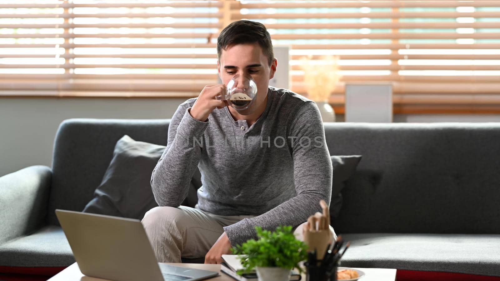 Young man drinking coffee and using computer laptop on couch.