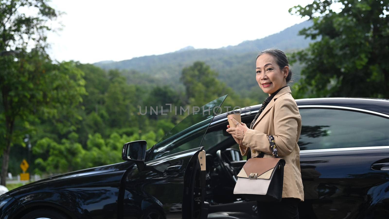 Mature business woman standing behind a car with opened door and smiling to camera.