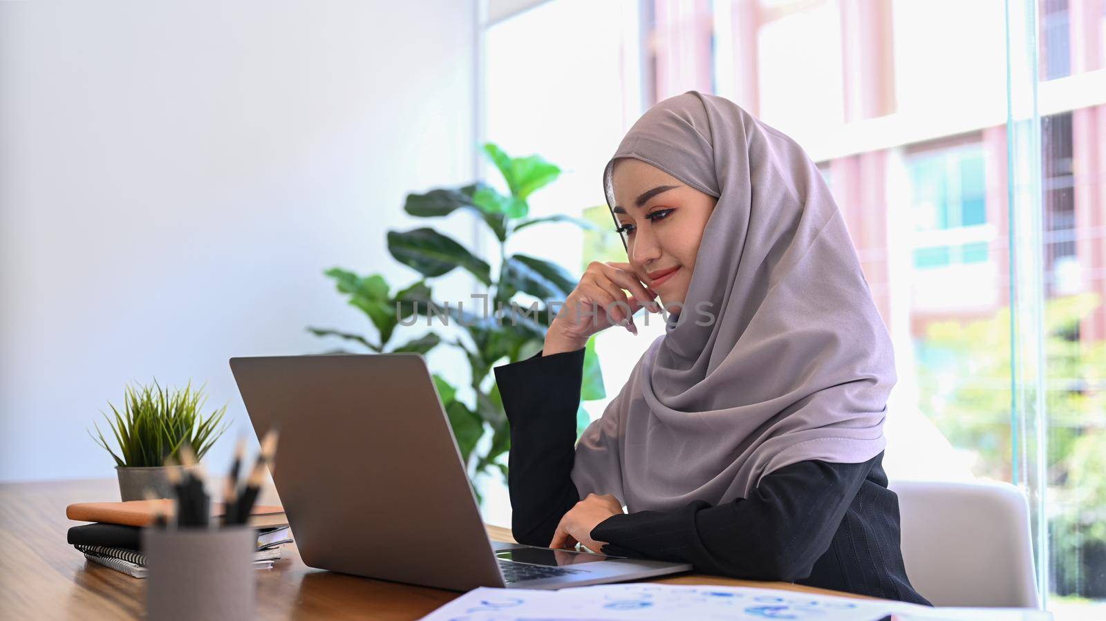 Beautiful young Muslim business woman in hijab sitting at modern office and watching online webinar on laptop by prathanchorruangsak