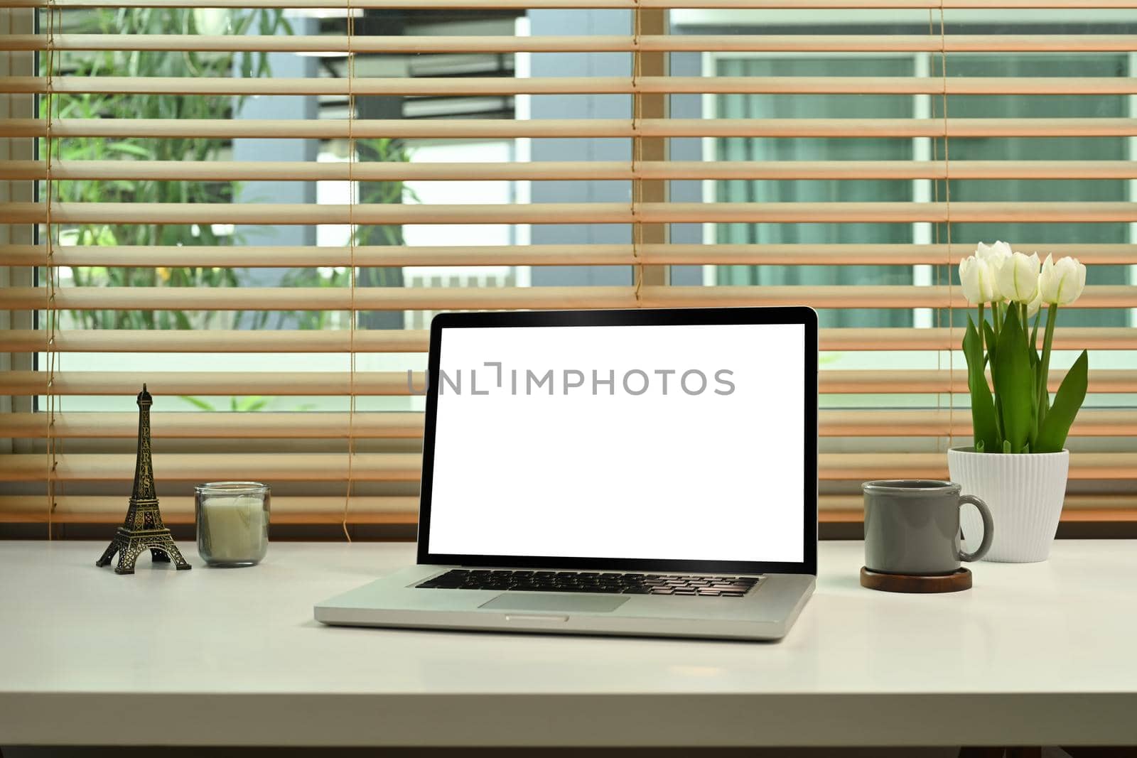 Computer laptop with empty display, coffee cup, candle and flower pot on white table.