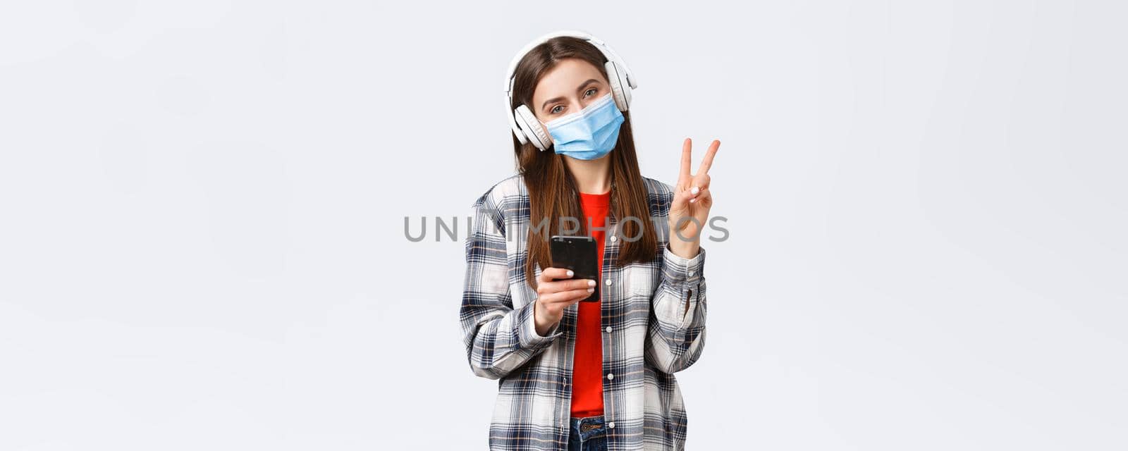 Social distancing, leisure and lifestyle on covid-19 outbreak, coronavirus concept. Cute silly teenage girl in medical mask and headphones, show peace sign, relaxing with music, hold mobile phone.
