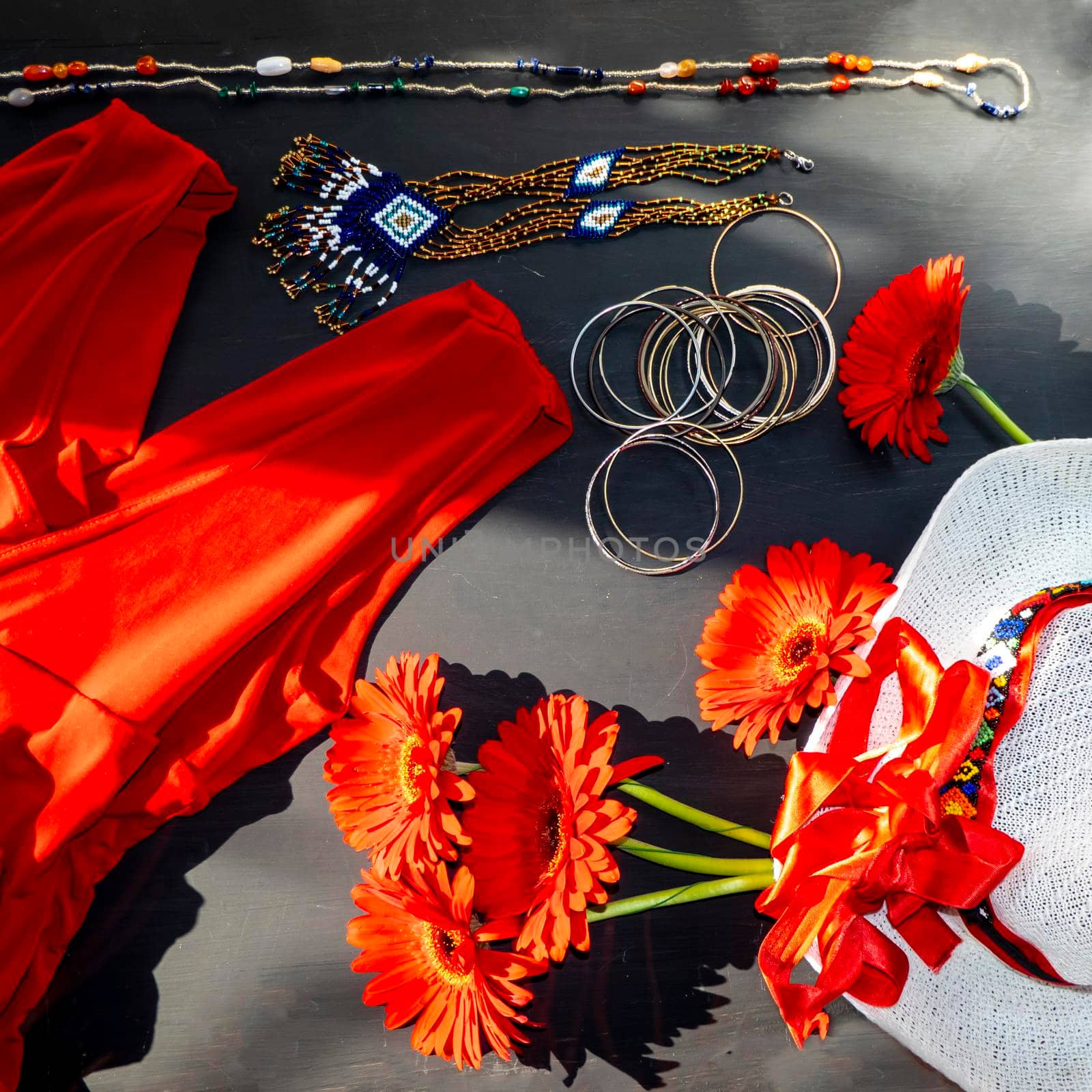 flat lay of a fashionable look from red and white accessories on a black background. Scarlet dress and white hat with a ribbon, jewelry made of beads and small stones, light jingling bracelets