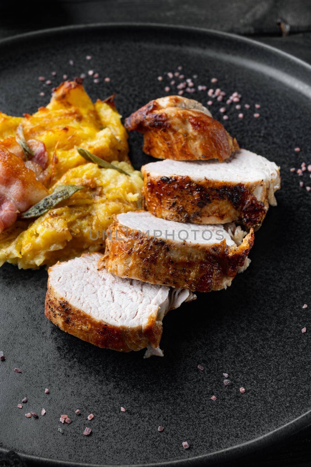 Roasted pork loin with mash potatoe gratin, sage and prosciutto set, on plate dish, on black wooden table background