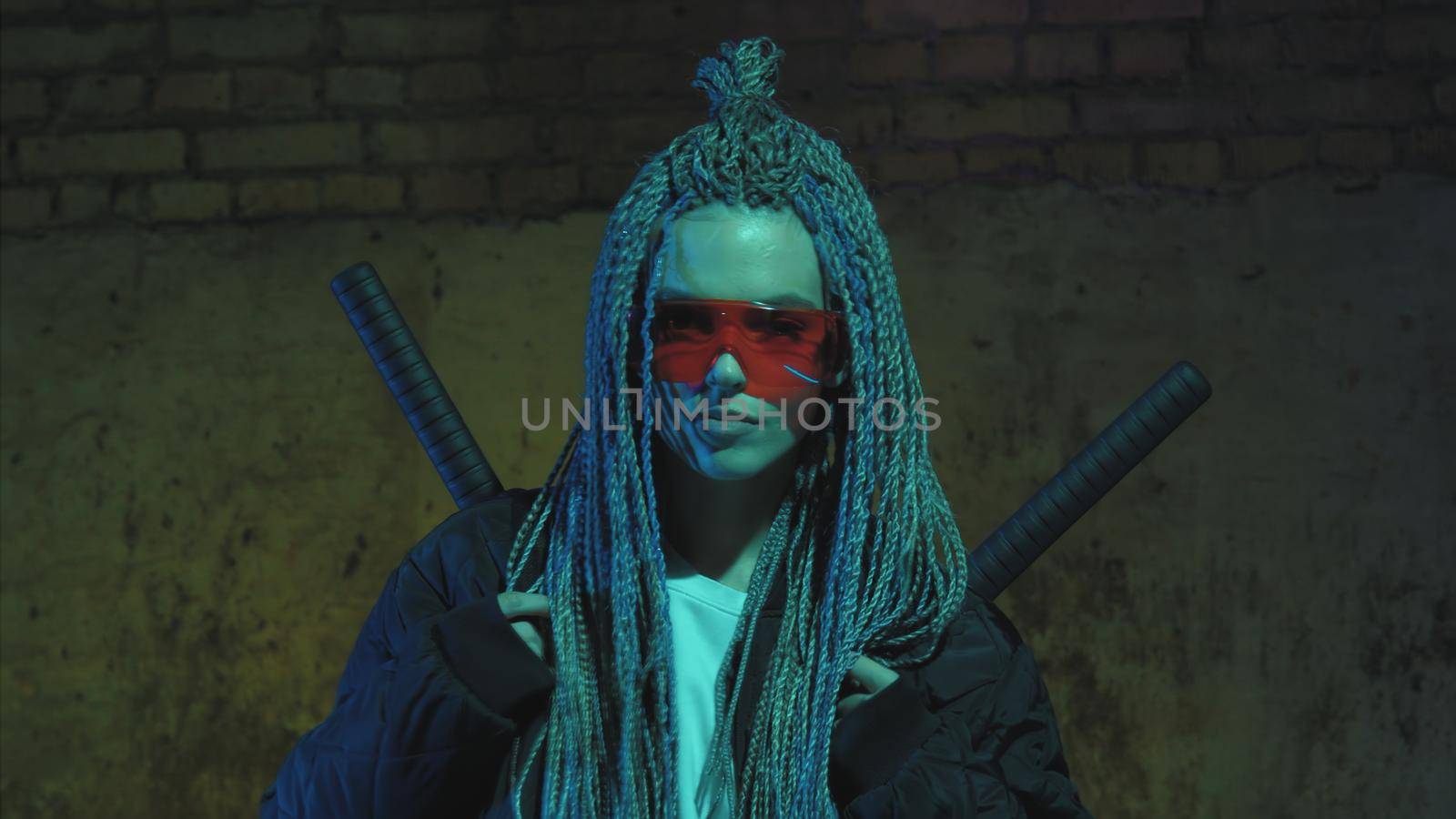 girl with dreadlocks and katanas in red glasses posing against a neon brick wall by studiodav