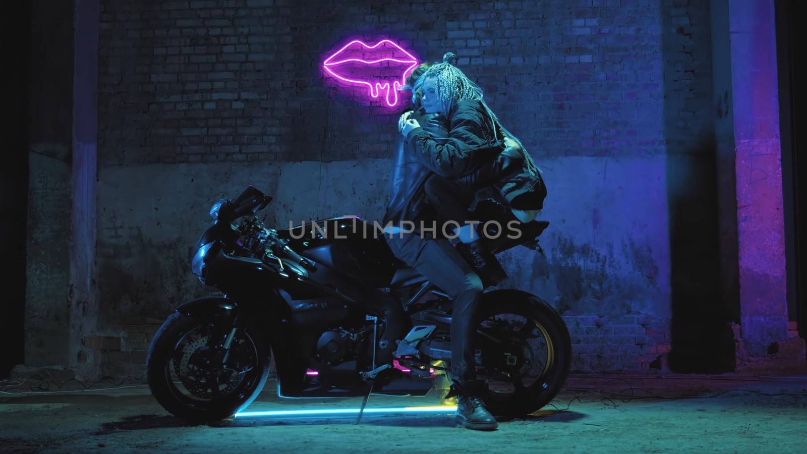 A girl in love and a guy are sitting on a super sport motorcycle flirting and hugging against the background of a neon sign by studiodav