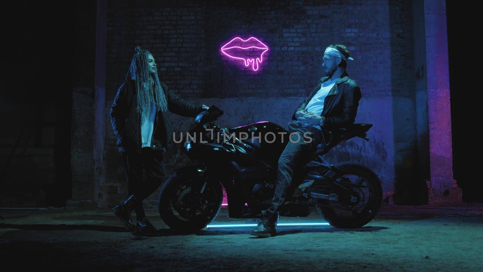 A guy on a super sport motorcycle stands talking to a girl against a pink neon sign by studiodav