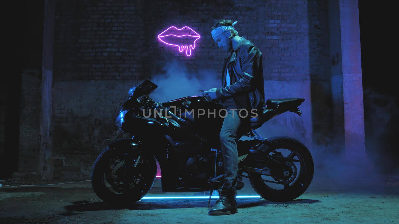 A man with a bandama sits on a sports motorcycle and texts on a mobile phone against a brick wall with a neon sign and smoke 4k