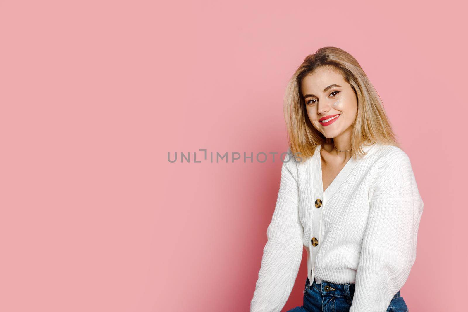 A cute fashion model in a white sweater smiles and looks at the camera against a pink background. Tenderness and youth.