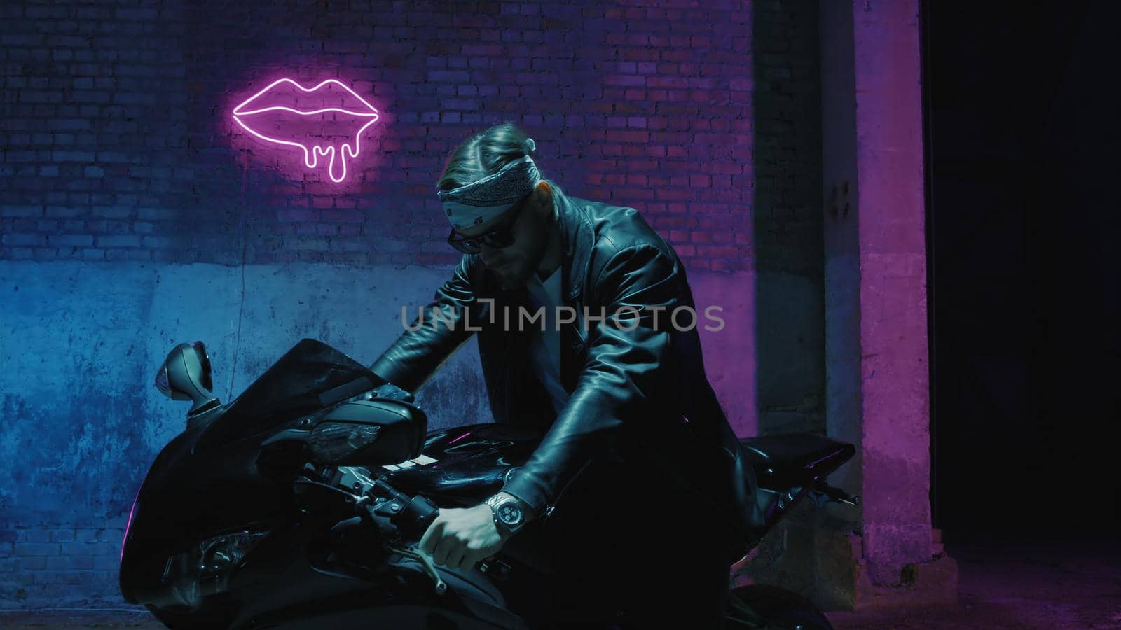 A man with a bandama sits on a sports motorcycle in sunglasses against a brick wall with a neon sign and smoke by studiodav
