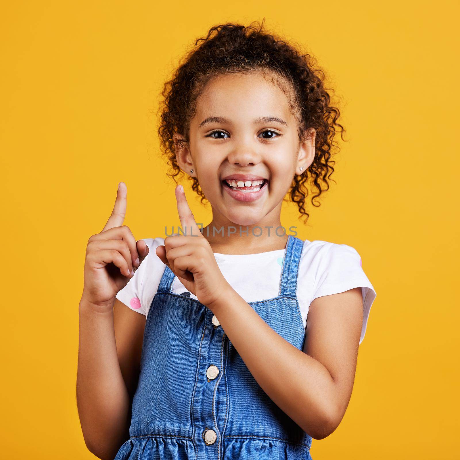 Studio portrait mixed race girl pointing upwards towards copyspace isolated against a yellow background. Cute hispanic child posing inside. Happy and cute kid showing or endorsing a company or product.