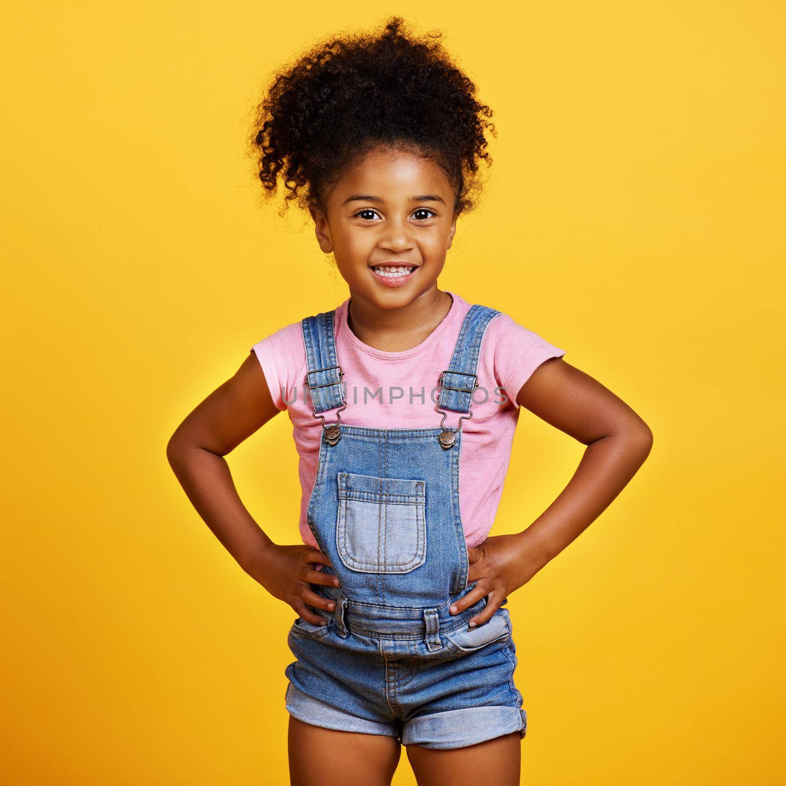 Studio portrait mixed race girl looking standing with her hands on her hips isolated against a yellow background. Cute hispanic child posing inside. Happy and cute kid smiling and looking confident.